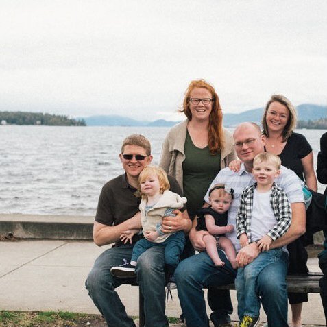 A group of six people, including three adults and three children, sitting and standing near a waterfront with mountains in the background.